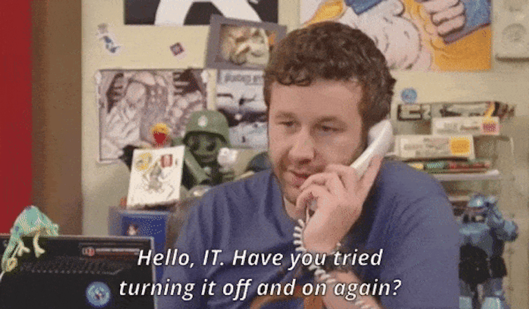 A man with curly hair, wearing a blue sweater, speaks on the phone in a cluttered office. He says, "Hello, IT. Have you tried turning it off and on again?.