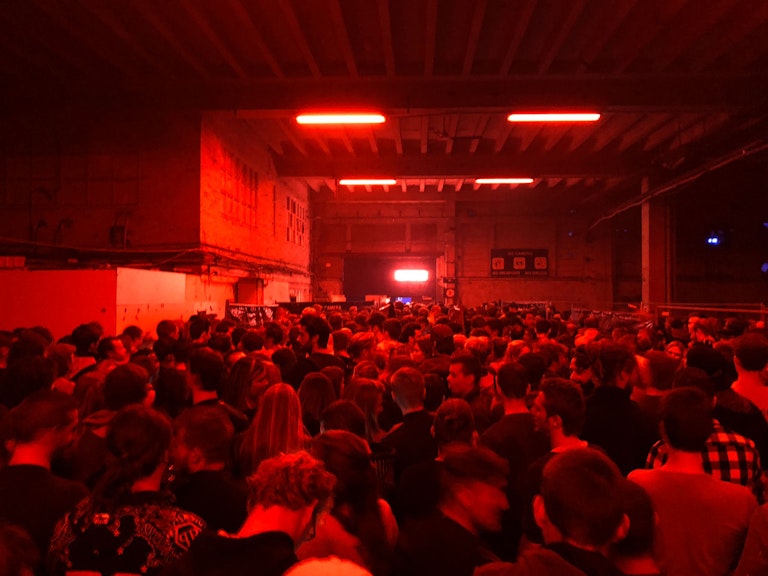 A crowded indoor concert with a sea of people illuminated by intense red lighting, creating a vibrant atmosphere.