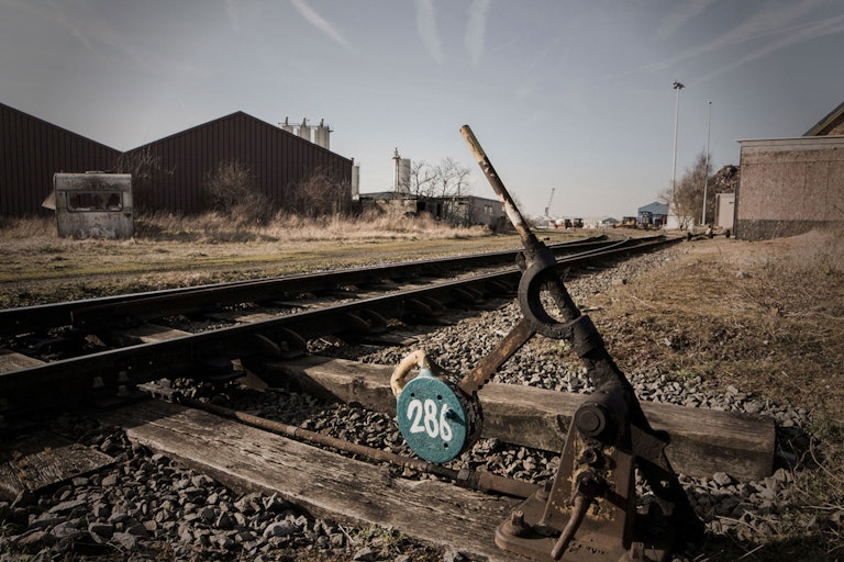 An old manual railroad switch lever, number 28, set against a backdrop of industrial buildings and rail tracks, under a clear sky.