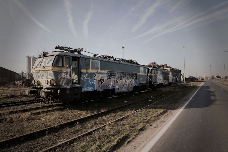 An old, graffiti-covered electric train sits abandoned on rusty tracks beside a road, under a cloudy sky. The setting has an abandoned industrial feel.