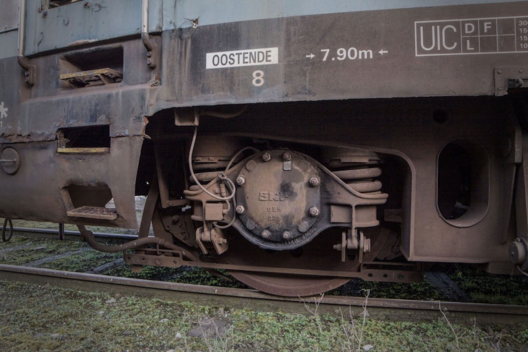 Close-up of the wheel and undercarriage of an old, rusty train car labeled "OOSTENDE" parked on railway tracks, showing details of its mechanical components and aging texture.