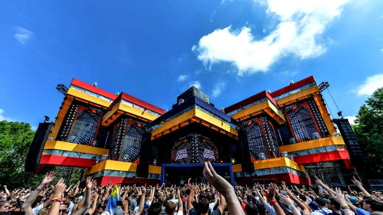 A vibrant music festival scene with a crowd of excited fans raising their hands under a blue sky, facing a large stage with bold red and yellow decorations.