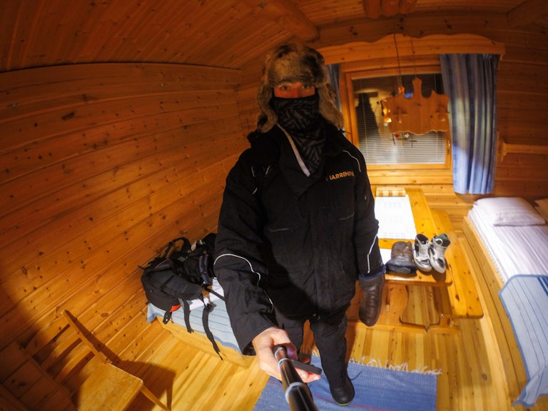 A person wearing winter gear and a fur hat stands inside a cozy wooden cabin, holding a camera for a selfie with a bed and window visible in the background.