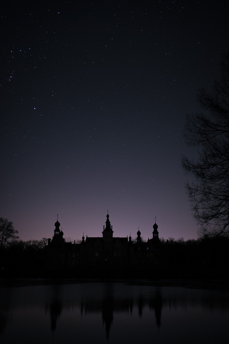 A silhouetted castle with multiple spires reflected in a still lake under a starry night sky, with trees visible on the right edge.