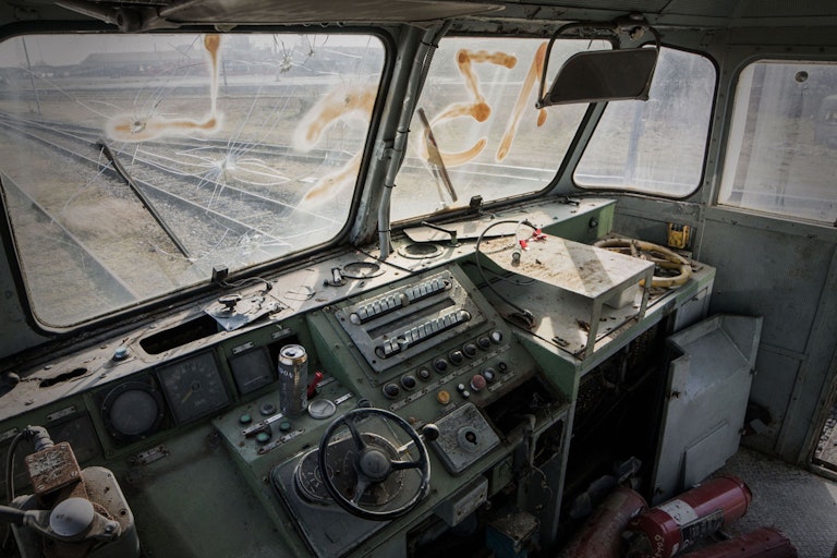 Interior of an abandoned train cab with weathered control panels, a cracked front window, scattered debris, and graffiti, giving a desolate and forsaken appearance.